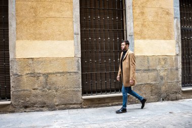 Şık bir ten rengi ceket giymiş genç bir adam sessiz ve dar bir sokakta geziniyor. Tarihi binada sessiz renkler ve zarif pencere ızgaraları yer alıyor. Atmosfer sakin ve yansıtıcı..