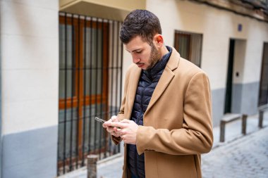 Şık ceketli genç bir adam, şık bir sokakta duran akıllı telefonuna odaklanmış durumda. Güneş, etrafındaki binaların büyüleyici mimarisini aydınlatan sıcak bir ışık yayar..