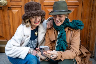 İki arkadaş dışarıda birbirine yakın oturuyor, gülümsüyor ve telefona bakarken bir anı paylaşıyorlar. Onların rahat şapkaları ve şık kıyafetleri serin güne sıcaklık katıyor..