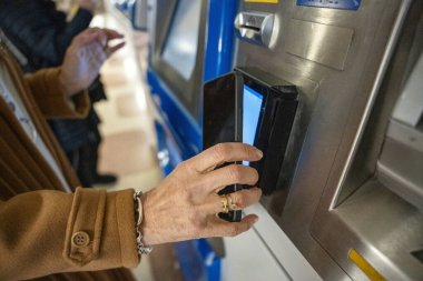 Şık bir ceketi olan biri, hareketli bir tren istasyonunda bilet makinesiyle etkileşim içindedir. Yerleşimciler yolculuklarına hazırlanırken ortam enerji ile titriyor..