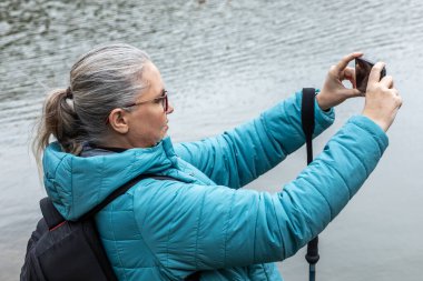 Deniz mavisi ceketli bir kadın suyun kenarında duruyor, neşeyle nehir kenarının sakin resimlerini çekiyor. Ağaçlar, güzelliklerle dolu huzurlu bir sonbahar gününü yansıtan altın renkler sergiliyor..