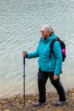 Soğuğa karşı sıcak giyinmiş bir kadın, sakin göl kıyısında yürüyor. Bir yürüyüş direği taşıyor, sakin bir sonbahar gününde sakin bir manzaranın tadını çıkarıyor..