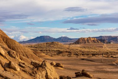 Uçsuz bucaksız kumlu arazi ve pürüzlü kaya oluşumları renkli bulutlara karşı yükseliyor..