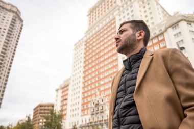 Genç bir adam yoğun şehir merkezinde düşünceli bir şekilde dikiliyor, yüksek binalarla çevrili. Gri bulutlar gökyüzünde sürüklenirken atmosfer dinamik hissediyor, değişen havayı ima ediyor..