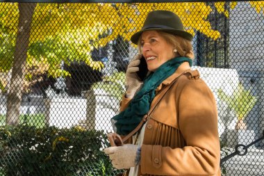 Parlak sonbahar renkleriyle çevrili şık bir ceket giymiş ve telefonunda sohbet eden gülümseyen bir kadın. Güneş ışığı altın yaprakların arasından süzülüp parkta neşeli bir atmosfer yaratır..