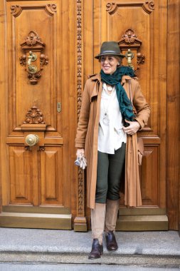 Modaya uygun bir kadın süslü ahşap kapıların yanında güvenle durur. O, rahat kahverengi bir ceket ve şapka giyerek zarafet yayar. Onun canlı eşarbı büyüleyici ortama renk katıyor..