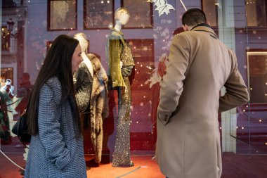 İki müşteri, gösterişli bir vitrin vitrininde sergilenen göz alıcı kıyafetlere hayran olmak için duruyor. Sahne, ışıldayan stiller ve bayram neşesiyle dolu kış alışverişinin heyecanını yakalıyor..