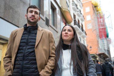 Kalabalık bir şehir caddesinde bir çift yakın yürür. Kış giysileri, mağazaları ve manzaraları keşfederken onları sıcak tutar. Gülüşleri serin havadaki neşeyi ve macerayı yansıtıyor..