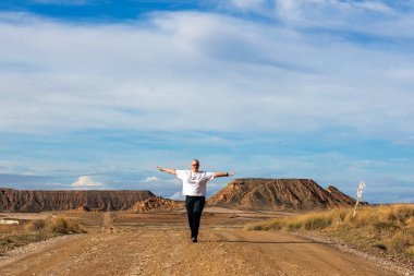 Bir insan tozlu bir yolda neşeli bir şekilde yürüyor, kolları açık, çarpıcı çöl manzarası ve parlak bir gökyüzünün altındaki görkemli kayalık tepelerle çevrili..