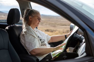 Bir kadın park halindeki arabasında oturuyor, haritaya derinden odaklanmış ve etrafını muazzam manzarayla çevreleyen. Yolculuğun tadını çıkarırken güneş ışığı onun derin derin ifadesine dokunur..
