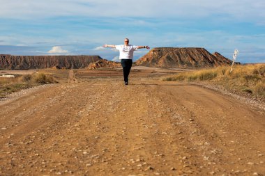 Titrek güneş, bir adamın virajlı toprak bir yol boyunca kollarını açarak yürüdüğü sakin bir sahneyi aydınlatıyor. Majestic Hills her iki tarafta da çarpıcı bir şekilde yükseliyor, doğal güzellikleri sessiz bir yerde sergiliyor..