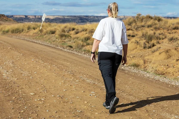 Bir kadın, altın çimenler ve kayalık oluşumlarla çevrili uzun toprak bir yolda rahat bir tempoda gezinir. Doğanın huzurlu huzurunun tadını çıkarırken güneş ışıl ışıl parlıyor..