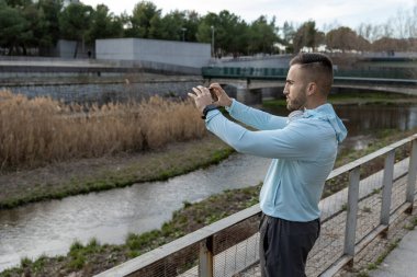 Sakin bir nehrin kenarındaki ahşap korkulukta duran bir adam sakin manzaranın fotoğraflarını çekiyor. Serin bir öğleden sonra, çevresini çevreleyen yeşillikler, huzur verici bir manzara yaratıyor..