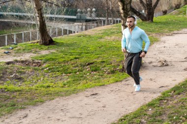 Açık mavi gömlekli bir adam sakin bir parkta ağaçlarla çevrili toprak bir patika boyunca koşuyor. Güneş doğar ve serin atmosfere sıcaklık katar. Temiz havanın tadını çıkarırken ayak altında çıtırdıyor..