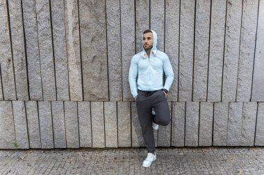 Genç bir adam hafif mavi spor kıyafetleri ve kulaklıklar içinde, çarpıcı taş bir duvara yaslanmış, hareketli bir şehir manzarasında bir anlık rahatlama keyfi yapıyor..