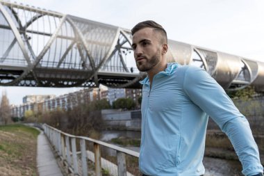 Açık mavi koşu tişörtlü genç bir adam nehir kenarındaki manzaralı bir yolda koşuyor. Yukarıdaki modern köprü kuleleri, sakin bir sabahta doğa ve şehir yaşamını harmanlıyor..