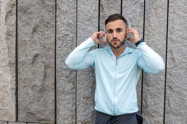 Kendini adamış bir fitness aşığı engebeli taş bir duvarın yanında duruyor açık mavi bir bluz ve kulaklık takıyor..