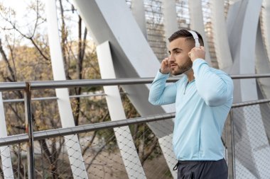Açık mavi spor bluzlu genç bir adam şık bir şehir köprüsünde duruyor, koşuya hazırlanırken kulaklıklarını ayarlıyor. Etrafındaki ağaçlar güneşin sıcaklığını görür ve canlı bir atmosfer yaratır..