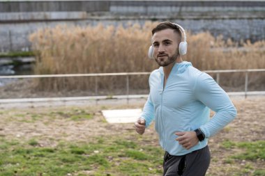 Bir adam dingin bir parkta, kulaklık ve açık mavi bir ceket takarak enerjik bir şekilde koşuyor. Uzun otlar suyun kenarında yavaşça sallanıyor. Egzersiz yapmaktan zevk alıyor..