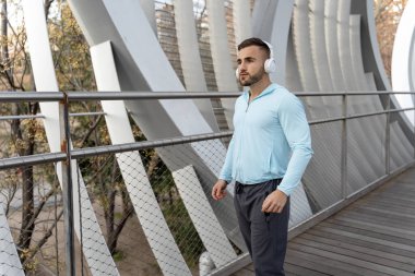 Genç bir adam parlak bir köprüde, kulaklık ve açık mavi bir bluzla çalışmaktan zevk alıyor. Güneş batıyor, koşusuna odaklanırken etrafındaki modern mimariyi vurguluyor..