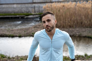 Genç bir adam sakin nehir kıyısında şık açık mavi ceketini sergileyerek güvenle duruyor. Güneş arka planda batıyor, uzun kamışlarla dolu huzurlu sahnenin üzerine ılık bir parıltı saçıyor..