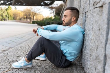 Genç bir adam, şehir parkında dokulu taş bir duvara yaslanmış bir şekilde huzur içinde oturuyor. Gökyüzü aydınlık ve berrak. Sıcak öğleden sonra güneşinin tadını çıkarıyor..