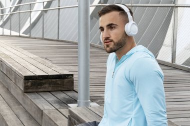 Kulaklık takan genç bir adam modern bir açık hava alanında ahşap merdivenlerde oturur. Etrafı gösterişli mimari ve sakin bir atmosferle çevriliyken müziği sever..