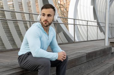 Bir adam ahşap bir bankta, kulaklık ve açık mavi bir ceket takarak huzur içinde oturuyor. Güneşli bir şehir parkındaki çevreye dikkatle bakıyor ve müziğinin tadını çıkarıyor..