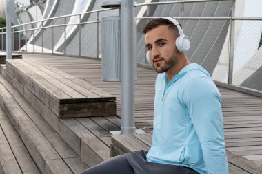 Genç bir adam ahşap bir güvertede büyük beyaz kulaklıklarla müzik dinliyor. Batan güneş, egzersizine ara verirken etrafında sıcak bir parıltı yaratıyor..