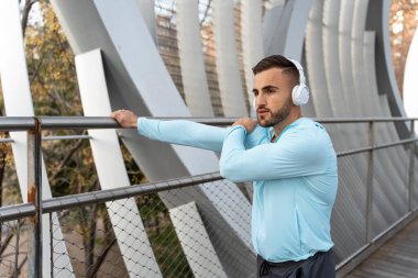 Açık mavi uzun kollu gömlekli genç bir adam modern bir köprüye uzanıyor. Beyaz kulaklıklarla müzik dinliyor, şehirde sabah sessizliğinin tadını çıkarıyor..