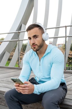 Şehir parkında ahşap bir platformda oturan genç bir adam. Açık mavi bir kapüşonlu ve büyük beyaz kulaklıklı, dikkatle akıllı telefonuna bakarken müziğinin tadını çıkarıyor..