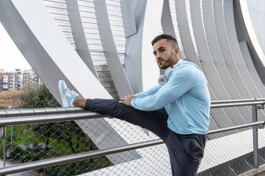 Spor kıyafetleri giymiş genç bir adam parlak bir korkuluğa yaslanırken bacağını esnetiyor. Şafak sökerken şehir manzarası yumuşak bir şekilde parlıyor, ferahlatıcı bir atmosfer yaratıyor..