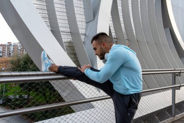 Açık mavi kapüşonlu bir adam gösterişli bir mimari arkaplana yaslanıyor. Dikkatle odaklanıyor, güneş batarken koşuya hazırlanıyor ve canlı bir atmosfer yaratıyor..