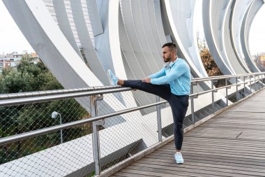 Sıradan bir atletik giysili bir adam, şık bir ahşap yolda bacağını korkuluklara dayar. Güneş ışıl ışıl parlıyor, etrafındaki eşsiz köprü dizaynını vurguluyor..