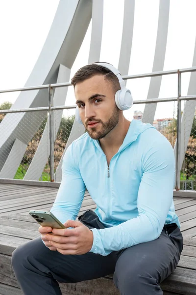 Şehir parkında ahşap bir platformda oturan genç bir adam. Açık mavi bir kapüşonlu ve büyük beyaz kulaklıklı, dikkatle akıllı telefonuna bakarken müziğinin tadını çıkarıyor..