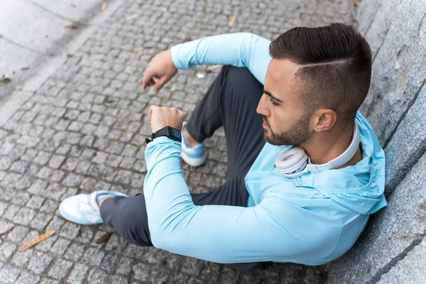 Açık mavi kapüşonlu genç bir adam taş bir duvara yaslanmış, spor saatine bakıyor, hareketli bir şehirde idman yaptıktan sonra dinleniyor..