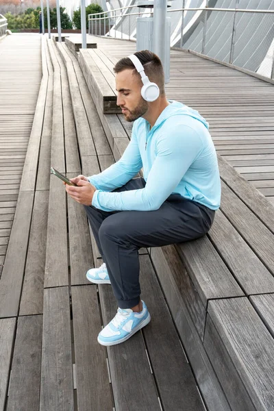 Genç bir adam açık terasta ahşap banklarda dinleniyor, akıllı telefonuna odaklanırken kulaklıkla müzik dinliyor. Güneş parlıyor, rahatlatıcı atmosferi güçlendiriyor..