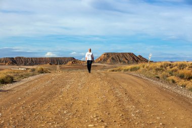 Tek bir figür, geniş bir çim ve kayalık tepelerle çevrili çakıllı bir yolda yürüyor. Güneş bu uzak ortamda huzurlu bir atmosfer yaratıyor..
