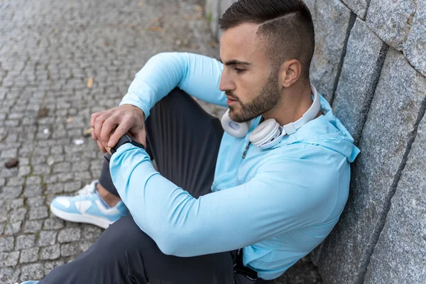 Formda genç bir adam kaldırım taşlı bir yolda durup rahat spor kıyafetleri giyerken saatine odaklanıyor. Öğleden sonra güneş ışığı, etrafında hafif gölgeler oluşturur..