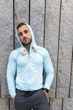 Genç adam, açık mavi kapüşonlu ve kulaklıklı, kendinden emin bir şekilde ayakta duruyor. Modern bir taş duvara karşı poz veriyor, şık bir şehir görüntüsü ve rahat bir tutum sergiliyor..