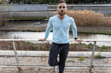Genç bir adam korkuluklara yaslanıyor, açık mavi bir bluz ve koyu bir pantolon giyiyor. Güneş sakin bir nehrin üzerinden yükselir ve çevreleyen yeşillik onun fitness rutinine huzur katar..