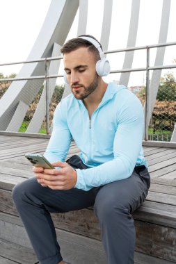 Kulaklıklı genç bir adam parktaki ahşap bir güvertede dinleniyor. Akıllı telefonuna odaklanıyor, yeşil gökyüzünün altında müziğin tadını çıkarıyor..