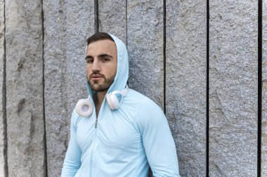 Genç bir adam, açık mavi kapüşonlu bir şeyle engebeli taş bir duvara karşı duruyor. İfadesi odaklanmış ve kulaklıkları boynunda duruyor. Şehir tarzında bir anı yakalıyor..