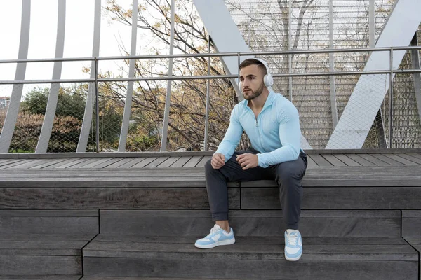 Genç bir adam, şehir parkında şık bir ahşap bankta, kulaklık takmış ve müziğinin tadını çıkararak rahatça oturuyor. Güneş batıyor, huzurlu anı güçlendiriyor..