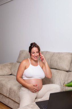 Hamile bir kadın yumuşak bir kanepede rahatça oturur, telefonda neşeyle sohbet eder ve nazikçe bebeğinin karnını şişirir. Yakınlarda açık bir dizüstü bilgisayar var, bu da sanal bir toplantı olduğunu gösteriyor..