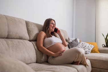 Hamile bir kadın, parlak oturma odasındaki yumuşak bir kanepede dinlenir. Telefonda konuşurken gülümsüyor, beklenti ve mutluluk anlarını kucaklıyor..