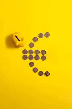 Yanında domuz kumbarası ve sarı arkaplan olan avro sikkelerinden yapılmış avro sembolünün üst görünümü