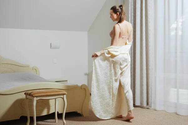 Bonita joven en el dormitorio quitándose peignoir de seda cerca de la cama —  Fotos de Stock