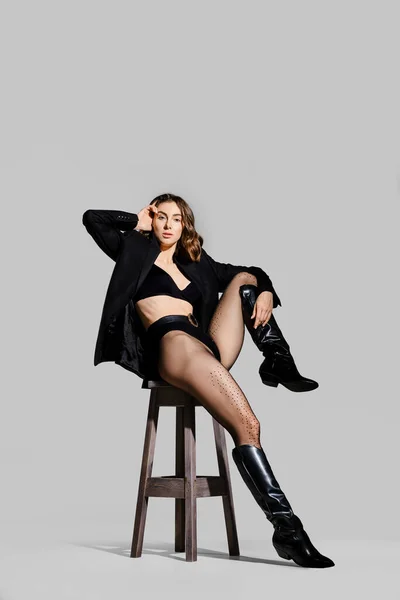 女性でBlazerと革のブーツ座っています上の椅子でスタジオとストレッチ彼女の足 — ストック写真