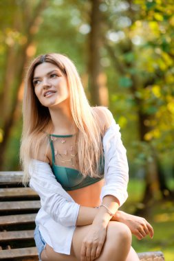 Yaz parkında güneş ışığı altında bankta oturan tatlı kız. O, beline bağlı kot şort ve beyaz gömlek giyiyor..